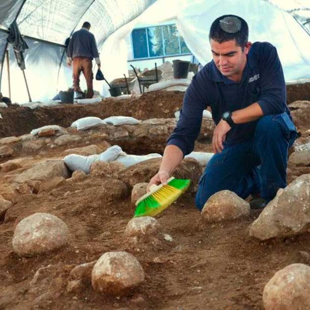 El director de excavación de IAA, Kfir Arbiv, limpia una piedra balista en el sitio de excavación del Complejo Ruso, que dice que fue arrojada por máquinas de asedio romanas que disparaban desde el barrio de Nahalat Hashiva en Jerusalén, a unos cientos de metros (yardas) al sur de donde se encontraron los proyectiles de piedra. (Yoli Schwartz/Autoridad de Antigüedades de Israel)