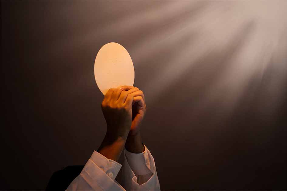 Hands of priest raise sacramental bread or the Eucharist under light. Source: Creativa Images/Adobe Stock