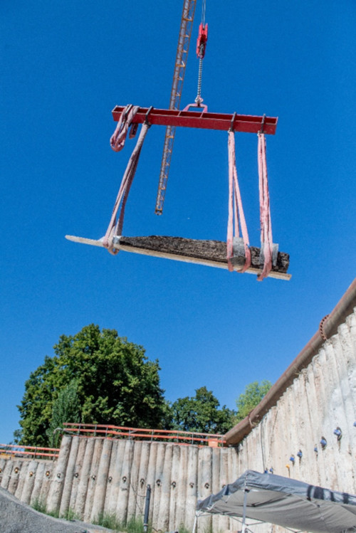 An oak beam is raised from the excavation pit by crane.