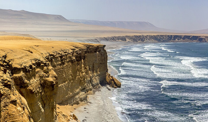 A sweeping aerial photograph of the dramatic golden cliffs and Pacific Ocean coastline at the Paracas National Reserve in Ica, Peru
