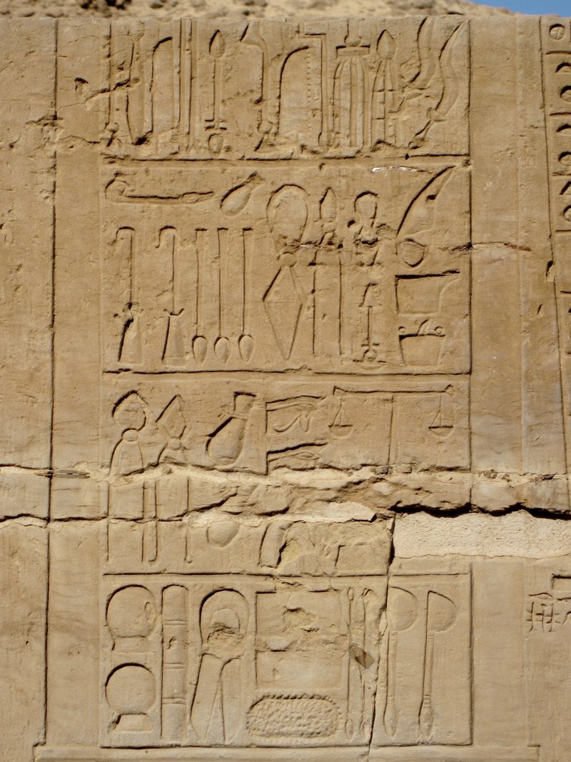 A carved temple wall showing rows of instruments including blades, hooks and containers beside hieroglyphs