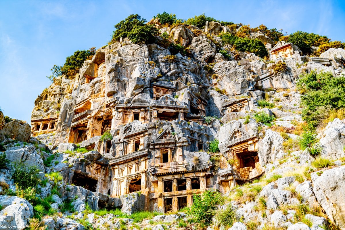 Temple fronted tombs carved into a rocky cliff face under a blue sky
