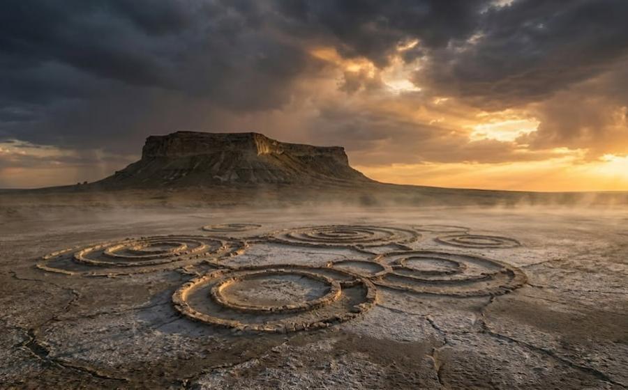 A haunting plateau rises over the cracked, salt-crusted desert. Mysterious circles etched into the ground hint at ancient secrets. Dramatic light and low mist shroud the landscape in enigma.