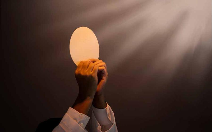 Hands of priest raise sacramental bread or the Eucharist under light. Source: Creativa Images/Adobe Stock