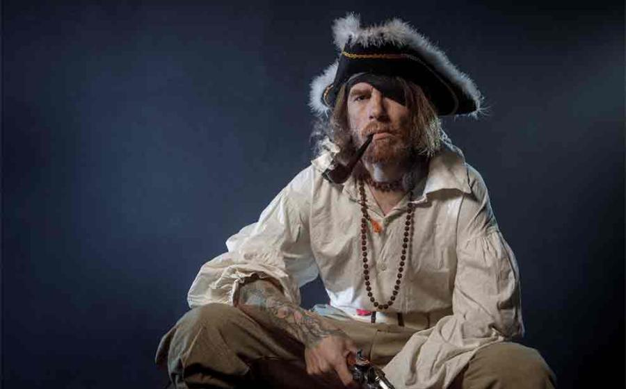 Image representing pirate in suit with gun and saber sitting on barrel. Source: Fotokvadrat/Adobe Stock