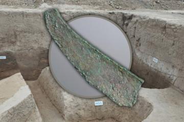 A broken bronze dagger-axe from the Jiangliu burial site, Shaanxi Province, with background of the excavation.