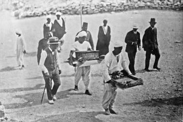 Black and white press photo of men in hats walking beside Egyptian workers who carry trays of objects across the valley floor