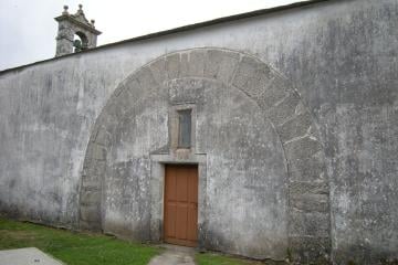 The whitewashed south wall of a small stone church, with a large round arch of stone blocks set into the wall around a wooden door