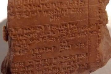 A brown baked clay tablet covered in rows of small wedge shaped cuneiform signs, displayed against a dark background