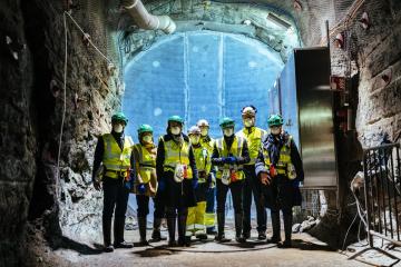 Workers in hard hats and high visibility vests stand in a rock tunnel deep underground at the Onkalo repository