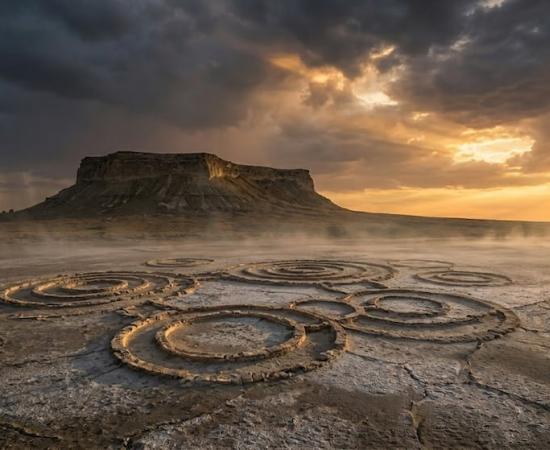 A haunting plateau rises over the cracked, salt-crusted desert. Mysterious circles etched into the ground hint at ancient secrets. Dramatic light and low mist shroud the landscape in enigma.