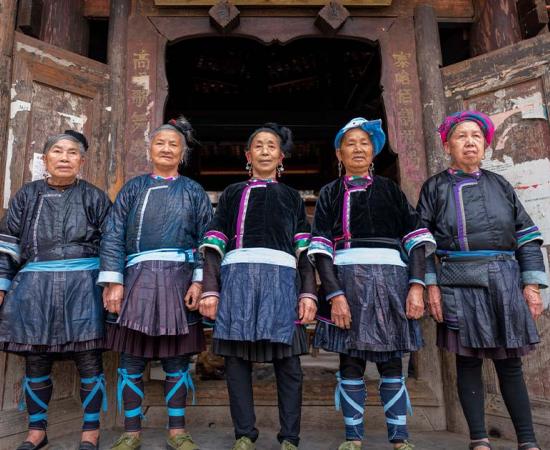 A singing group of 5 ethnic Kam women stand in front of the wooden drum tower in Xiaohuang village, Congjiang county, Guizhou, China. Source: imphilip / Adobe Stock.