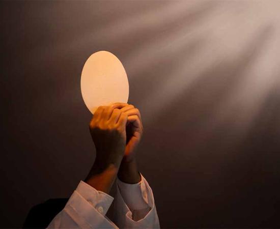 Hands of priest raise sacramental bread or the Eucharist under light. Source: Creativa Images/Adobe Stock