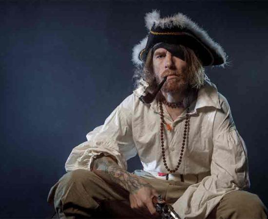 Image representing pirate in suit with gun and saber sitting on barrel. Source: Fotokvadrat/Adobe Stock