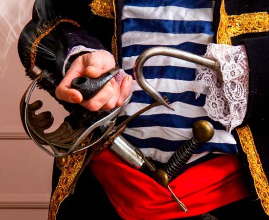 Close-up representation of an armed pirate captain in a frock coat and a vest with a sword, dagger and a hook, smoking a pipe. Source: sharpner/Adobe Stock
