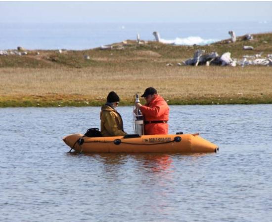 Sampling crew collecting a sediment core at PaJs-13, a Thule-Inuit site on Somerset Island, Nunavut, with the remnants of whalebone houses visible in the background.	Source: Jules Blais/University of Ottawa