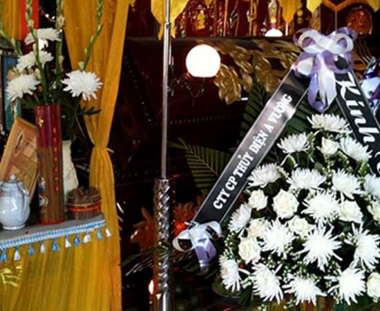Decorations placed around a coffin at a home funeral in Da Nang, Vietnam. At left, placed in front of the coffin, is an altar featuring a framed photo of the deceased and a pot for offering joss sticks. At right are a number of flower bouquets with attached condolences, and in the background are a number of vertical banners, also offering condolences.