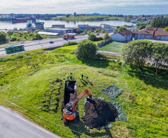 Aerial view of the Grønhaug excavation, with the mound and trench beside the Karmsundet landscape.