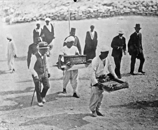 Black and white press photo of men in hats walking beside Egyptian workers who carry trays of objects across the valley floor