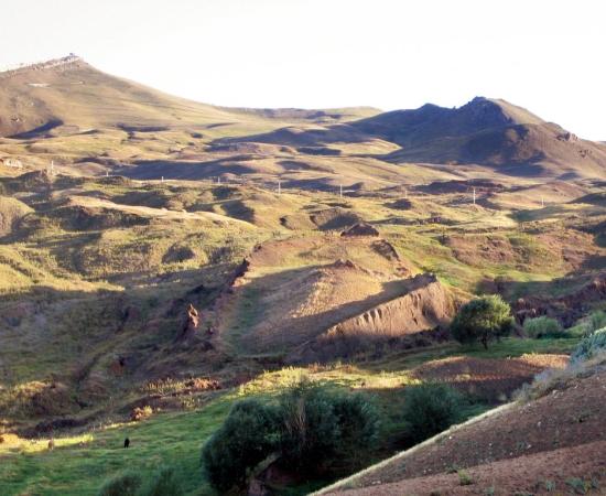 Grassy hillside valley in eastern Turkey with a long, raised, boat-shaped earth formation in the center and mountains behind