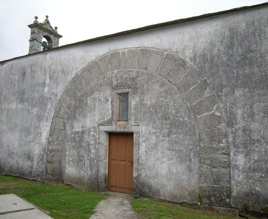 The whitewashed south wall of a small stone church, with a large round arch of stone blocks set into the wall around a wooden door