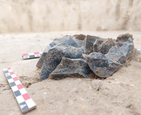 A small heap of dark flint pieces lying in pale sand at an excavation, with red and white scale bars