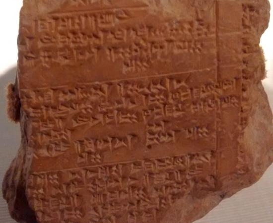 A brown baked clay tablet covered in rows of small wedge shaped cuneiform signs, displayed against a dark background
