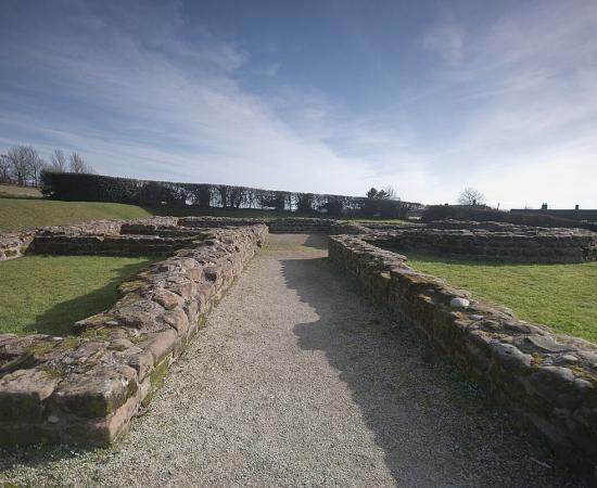 Low stone foundations of a Roman building laid out on grass under a blue sky, with a gravel path running between two walls