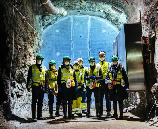 Workers in hard hats and high visibility vests stand in a rock tunnel deep underground at the Onkalo repository