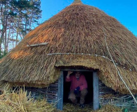 A second reconstructed roundhouse has been built by University of Dublin Centre for Experimental Archaeology students, after the first was subject to arson. Dr. Aidan O’Sullivan, crouched in the entryway of the roundhouse his students built. Source: UCD Centre for Experimental Archaeology