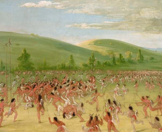 Hundreds of Choctaw players holding sticks chase a ball across a wide green field below rolling hills
