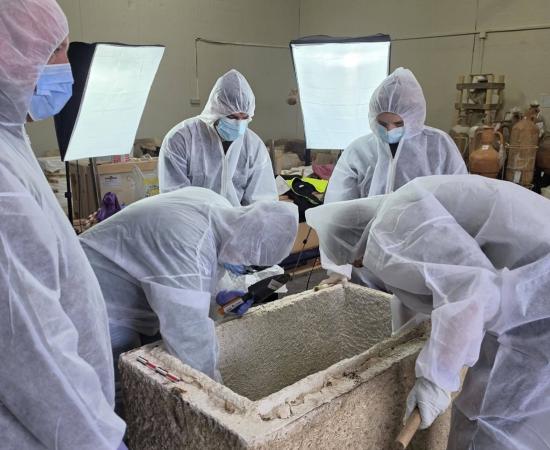 Museum specialists in white protective suits leaning over an open stone sarcophagus