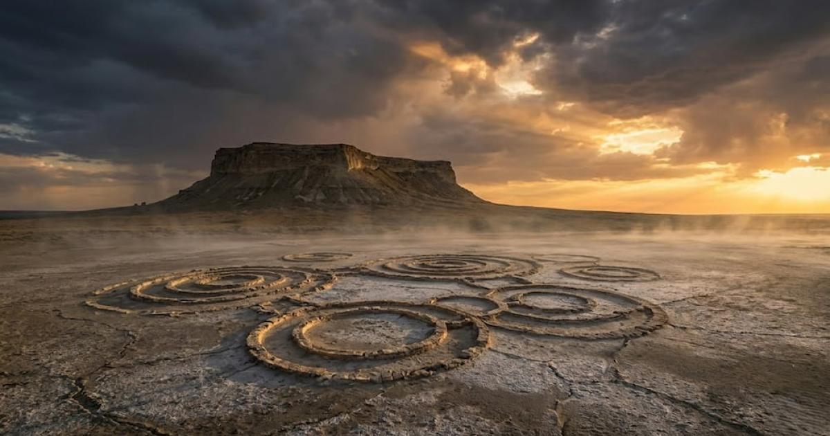 A haunting plateau rises over the cracked, salt-crusted desert. Mysterious circles etched into the ground hint at ancient secrets. Dramatic light and low mist shroud the landscape in enigma.