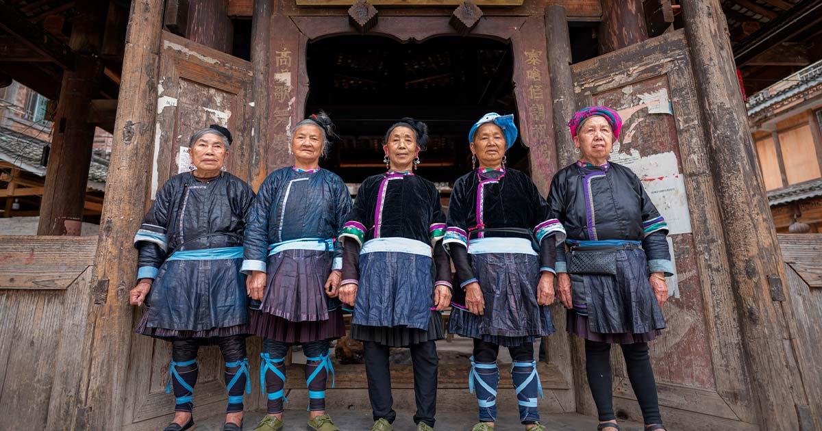 A singing group of 5 ethnic Kam women stand in front of the wooden drum tower in Xiaohuang village, Congjiang county, Guizhou, China. Source: imphilip / Adobe Stock.