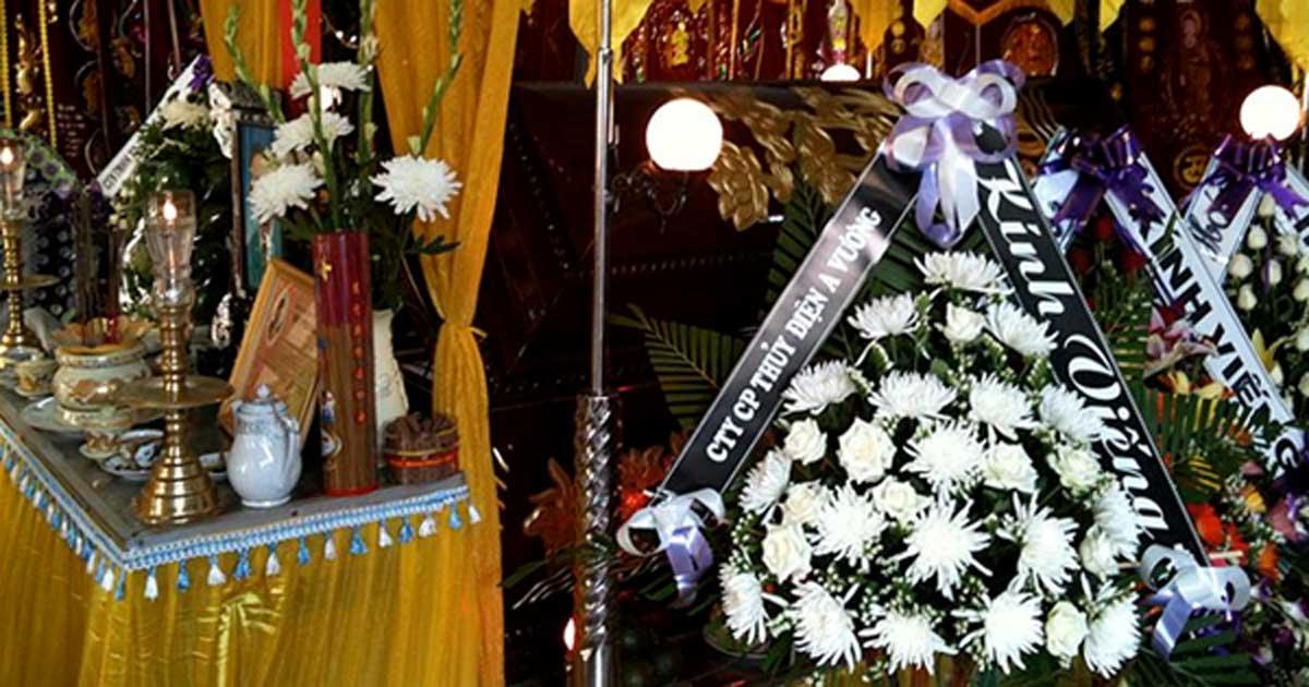 Decorations placed around a coffin at a home funeral in Da Nang, Vietnam. At left, placed in front of the coffin, is an altar featuring a framed photo of the deceased and a pot for offering joss sticks. At right are a number of flower bouquets with attached condolences, and in the background are a number of vertical banners, also offering condolences.