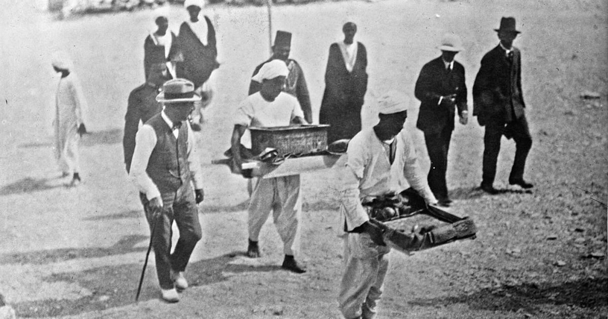 Black and white press photo of men in hats walking beside Egyptian workers who carry trays of objects across the valley floor