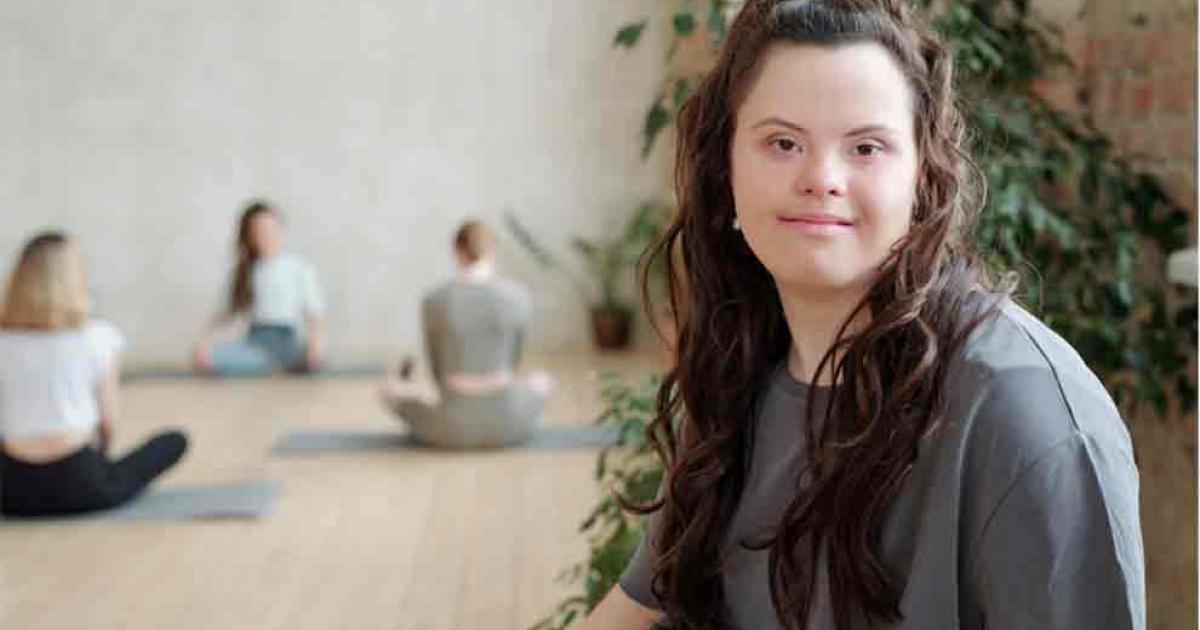 Modern day woman with Down syndrome looking at you while group of girls practicing yoga in gym. Source: pressmaster/Adobe Stock