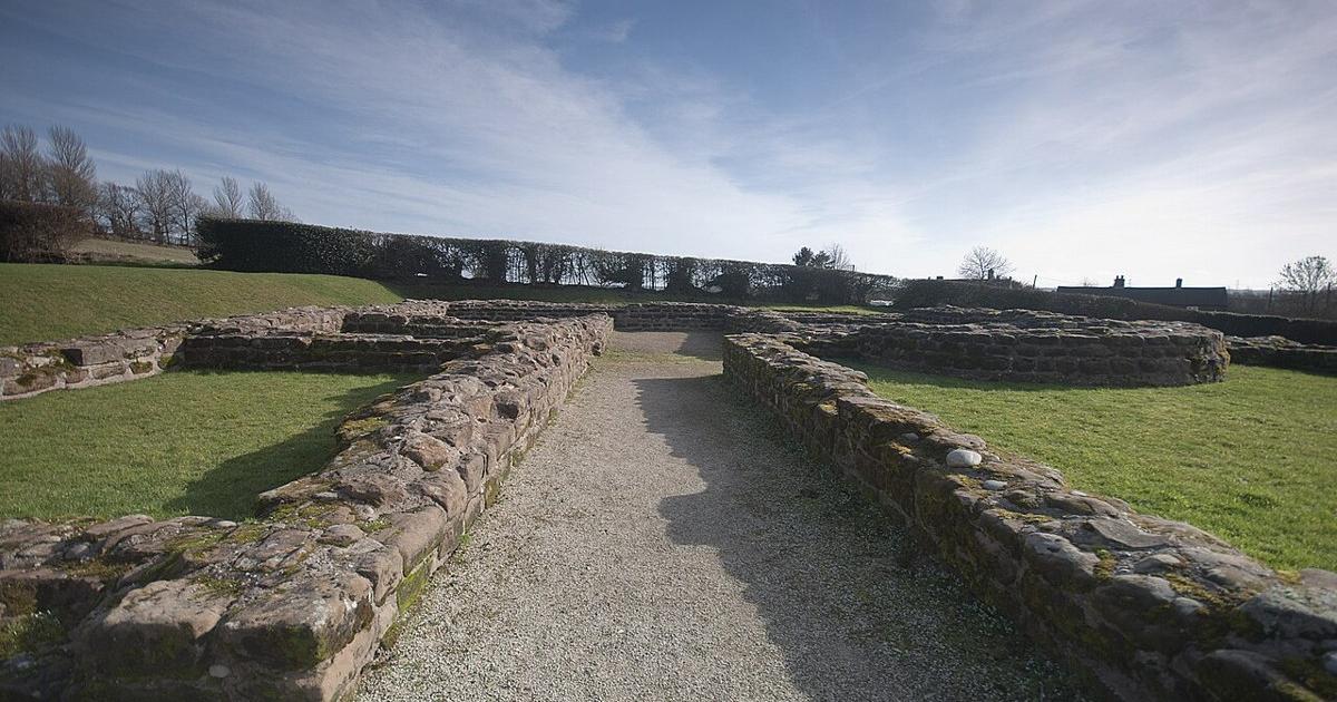 Low stone foundations of a Roman building laid out on grass under a blue sky, with a gravel path running between two walls