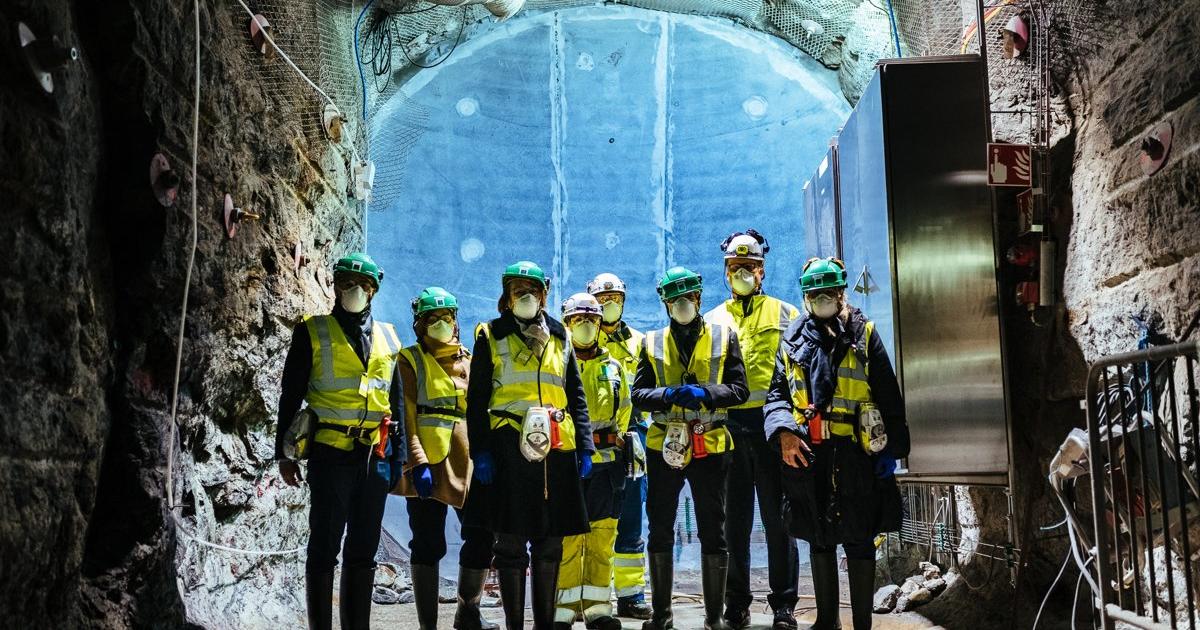 Workers in hard hats and high visibility vests stand in a rock tunnel deep underground at the Onkalo repository