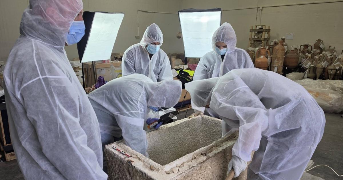 Museum specialists in white protective suits leaning over an open stone sarcophagus