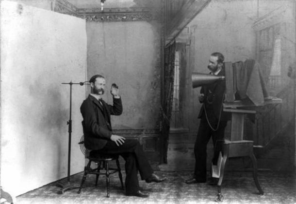 A composite photograph from 1893 showing a photographic studio interior. One man is seated on a stool near an adjustable clamp to hold his head steady during a long portrait exposure. The second man, standing next to a large view camera, looks like the person being photographed. (Public Domain)
