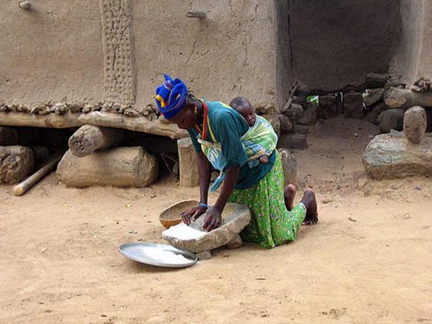 Woman grinding flour in front of traditional Dogon grain storage sheds. 
