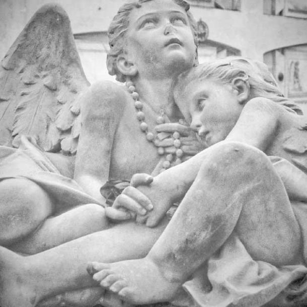 A child curls up in the arms of a young angel on top of a tomb in Staglieno Cemetery (lnm / Adobe Stock)