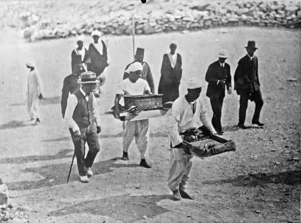 Black and white press photo of men in hats walking beside Egyptian workers who carry trays of objects across the valley floor