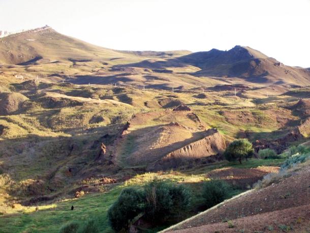Grassy hillside valley in eastern Turkey with a long, raised, boat-shaped earth formation in the center and mountains behind