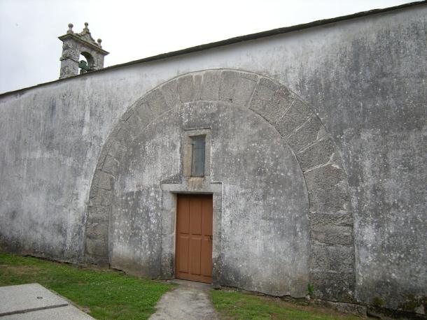 The whitewashed south wall of a small stone church, with a large round arch of stone blocks set into the wall around a wooden door