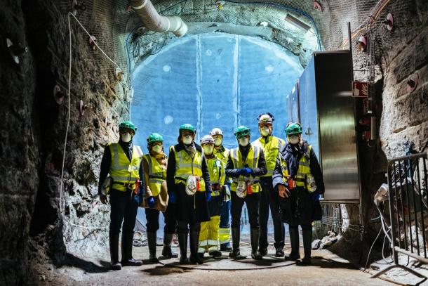 Workers in hard hats and high visibility vests stand in a rock tunnel deep underground at the Onkalo repository