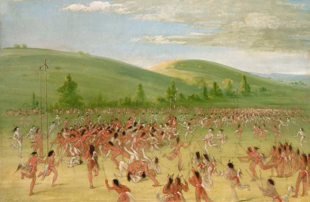 Hundreds of Choctaw players holding sticks chase a ball across a wide green field below rolling hills