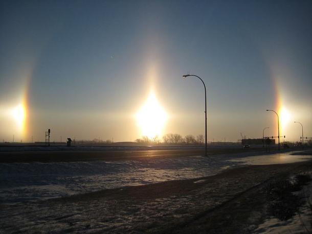 Very bright sun dogs in Fargo, North Dakota. Also visible are parts of the 22° halo (the arcs passing through each sundog), a sun pillar (the vertical line) and the parhelic circle (the horizontal line). (Public Domain)