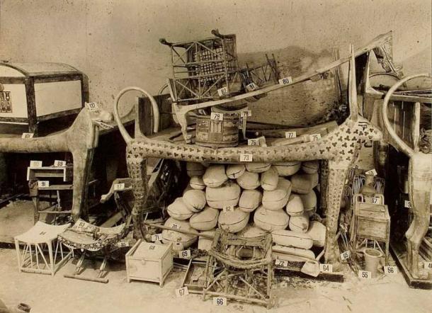 Cow-headed couch in the Antechamber of Tutankhamun’s tomb with many objects stacked on top. The packages underneath contained joints of meat. (Public Domain)